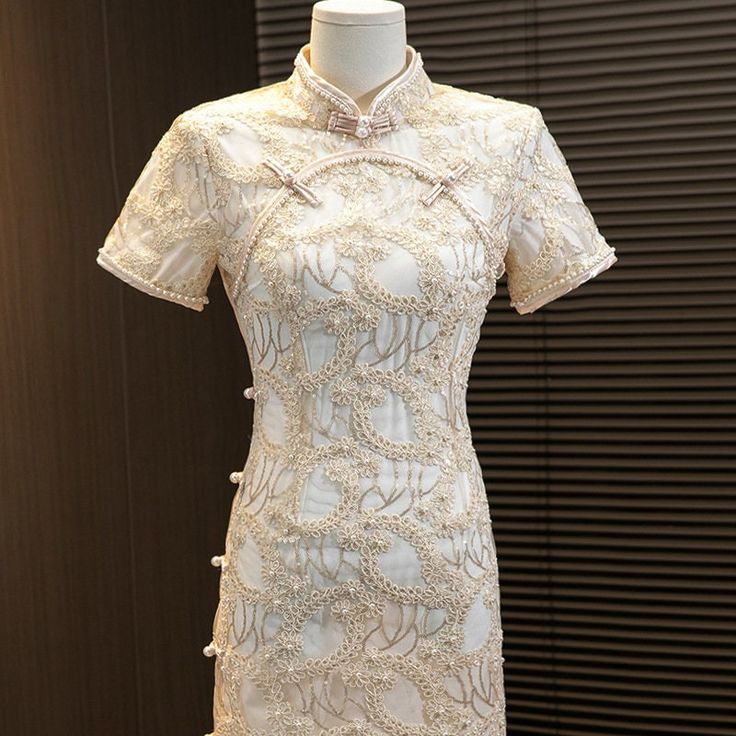 Introducing the stunning short sleeves white lace qipao with delicate floral appliques and faux pearls, a true masterpiece in traditional Chinese fashion. This gorgeous dress is crafted from high-quality lace material that feels soft and comfortable against the skin.The dress features intricate floral appliques that are intricately sewn onto the fabric, creating a beautiful and intricate design that is both feminine and elegant. Faux pearls are also incorporated into the design, adding a touch o Elegant Short Sleeve Embroidered Lace Dress, Traditional White Cheongsam With Stand Collar, Traditional Short Sleeve Fitted Cheongsam, Traditional Beige Lace Dress, Elegant White Cheongsam For Spring, White Embroidered Lace Dress With Short Sleeves, Elegant Lace Dress With Floral Embroidery And Short Sleeves, Elegant Embroidered Cheongsam For Spring, Traditional Short Sleeve Cheongsam For Wedding