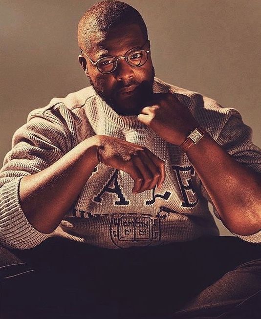 a man with glasses sitting on the floor in front of a wall wearing a sweater