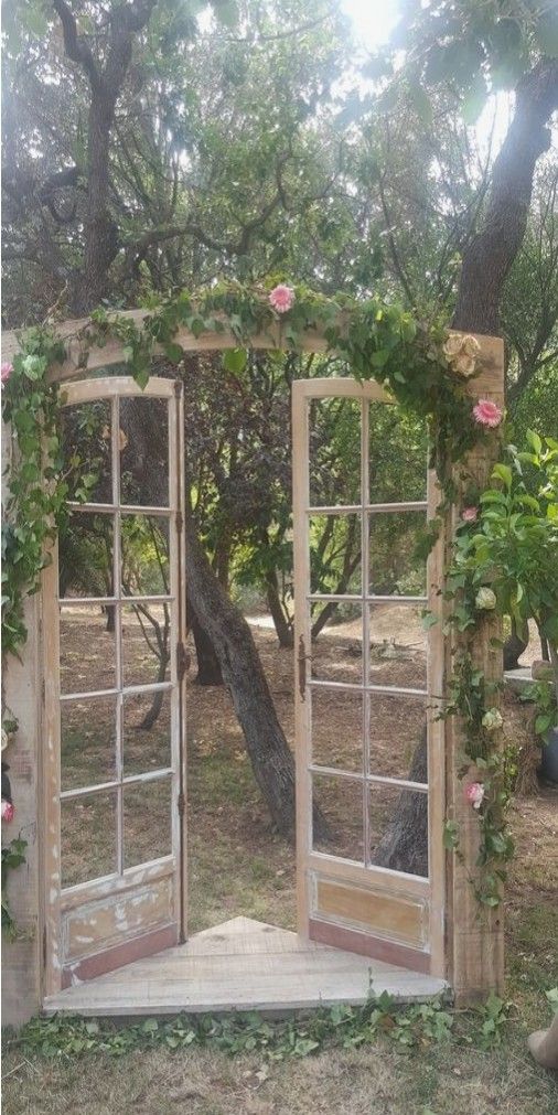 an open wooden door in the middle of a yard with flowers growing on it and trees around