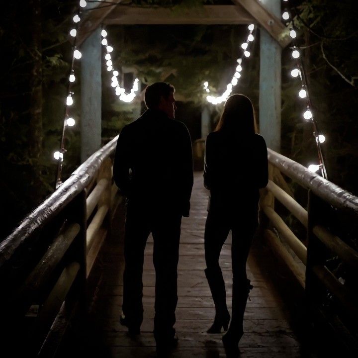 two people standing on a bridge at night with fairy lights strung all over the walkway