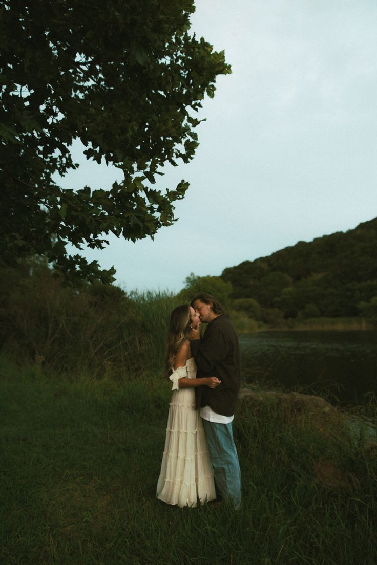 a man and woman standing next to each other in the grass near a lake at dusk