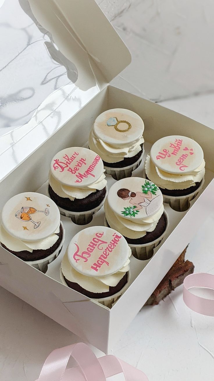 six decorated cupcakes in a white box with pink ribbons on the table next to it