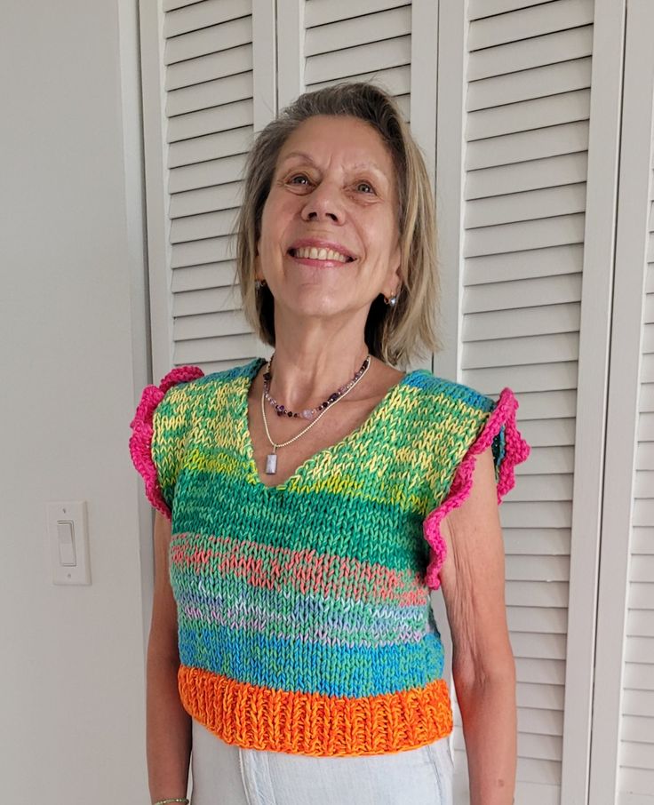 an older woman standing in front of shutters wearing a multicolored knitted top