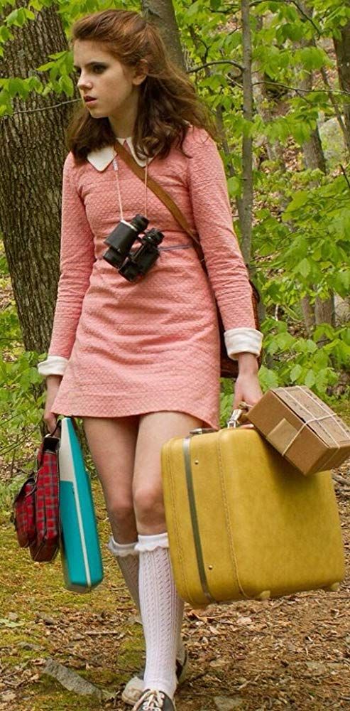 a woman in pink dress carrying two suitcases and a brown bag on her shoulder