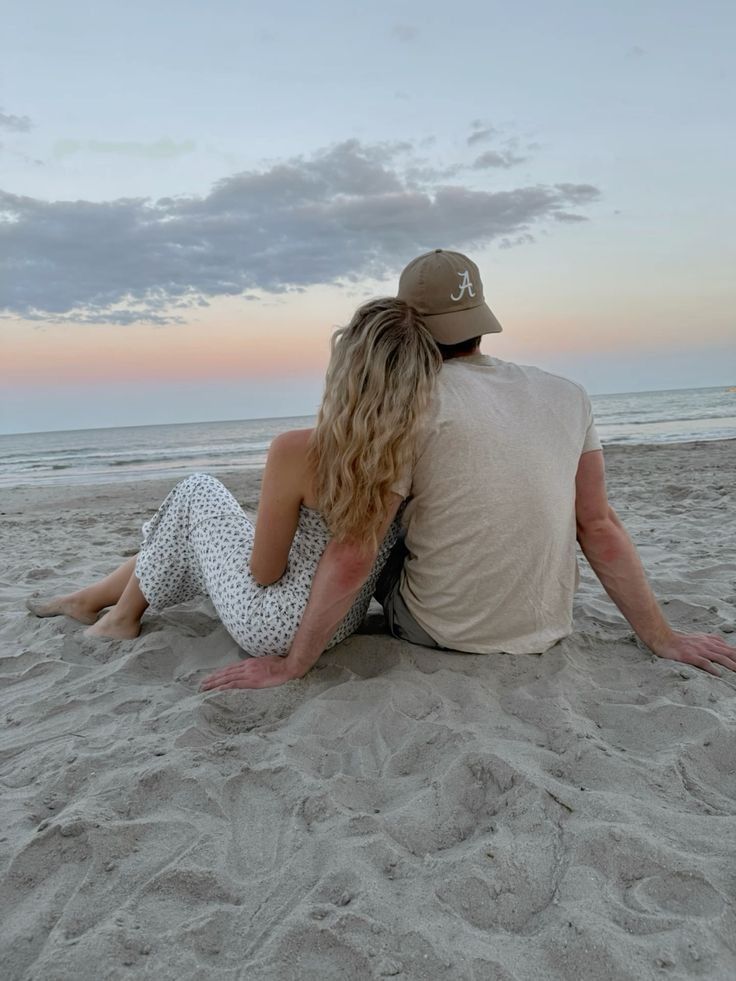 a man and woman sitting on the beach looking out at the ocean while the sun is setting