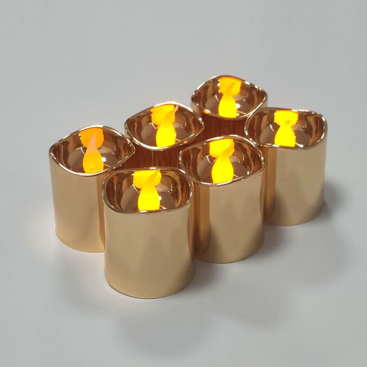 six gold cups with yellow candles in them on a gray surface, one is lit up and the other is empty