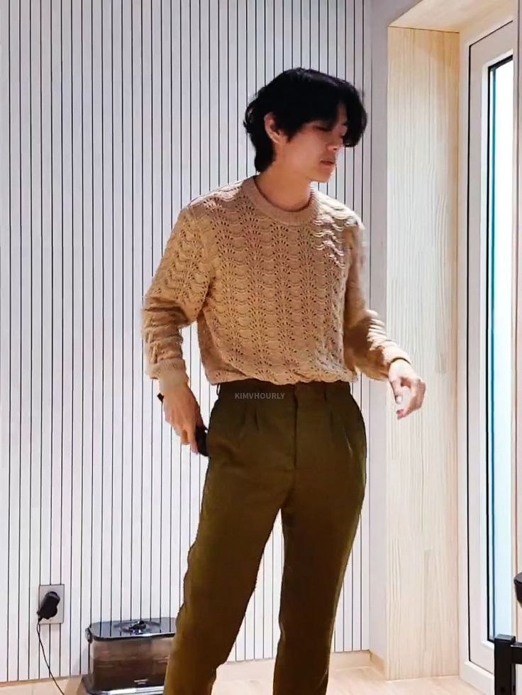 a young man standing in front of a window wearing brown pants and a beige sweater