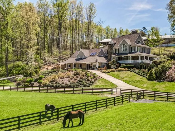 two horses graze in front of a large house on a hill with trees and grass