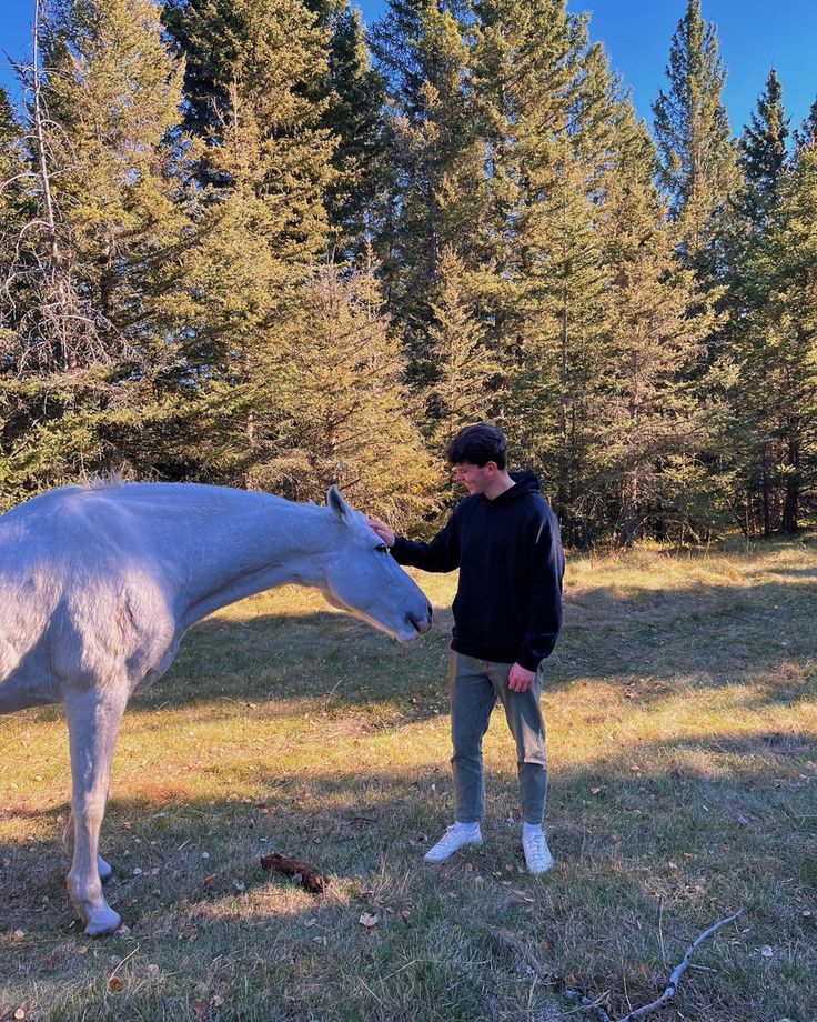 a man standing next to a white horse on top of a grass covered field with trees in the background