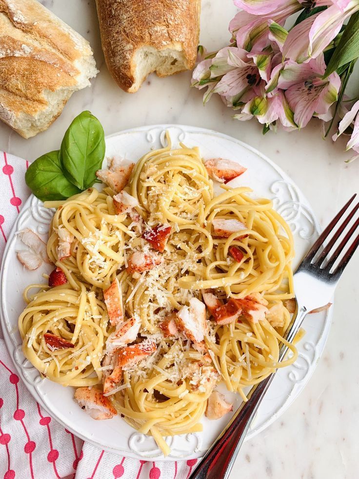 a white plate topped with pasta and meat next to bread on a tablecloth near flowers