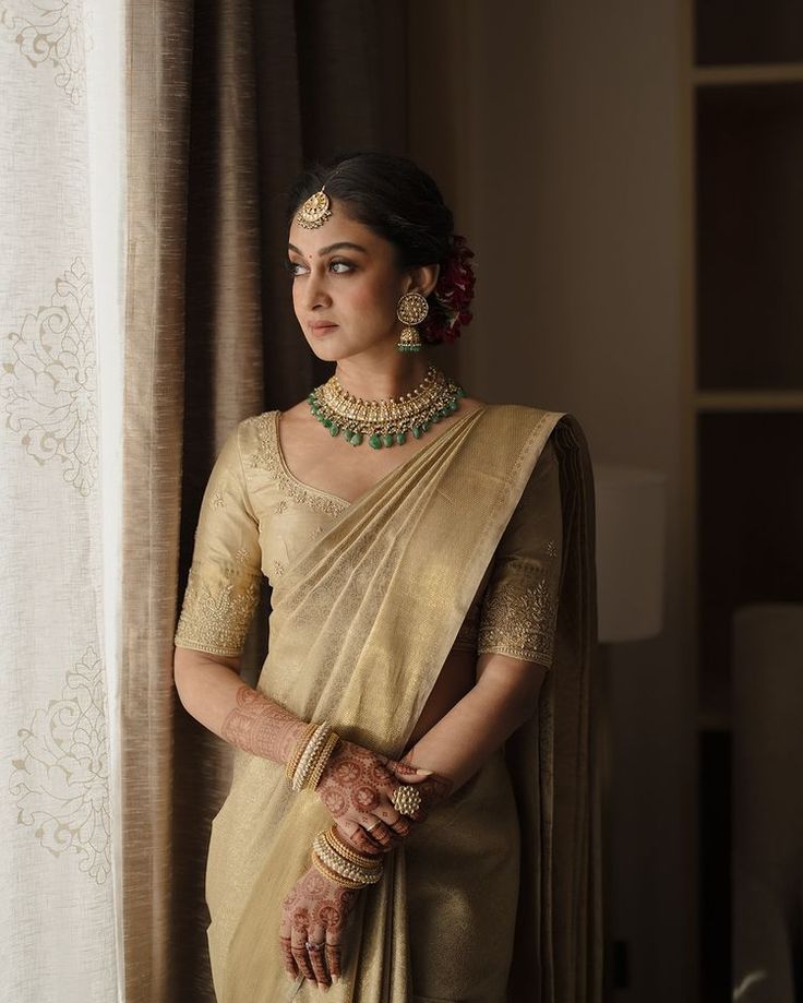 a woman in a beige sari with jewelry on her neck and hands behind her back