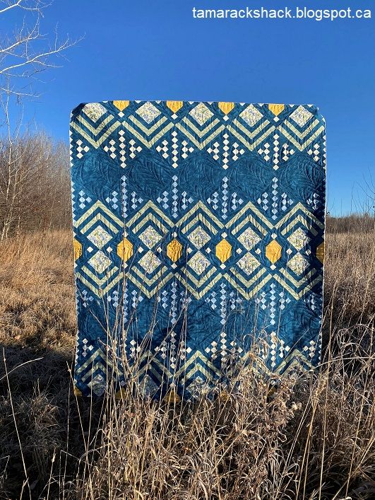 a blue and yellow quilt sitting in the middle of a dry grass field with trees