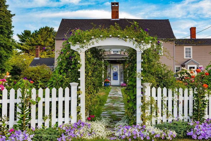 a white picket fence surrounded by flowers and greenery in front of a large house