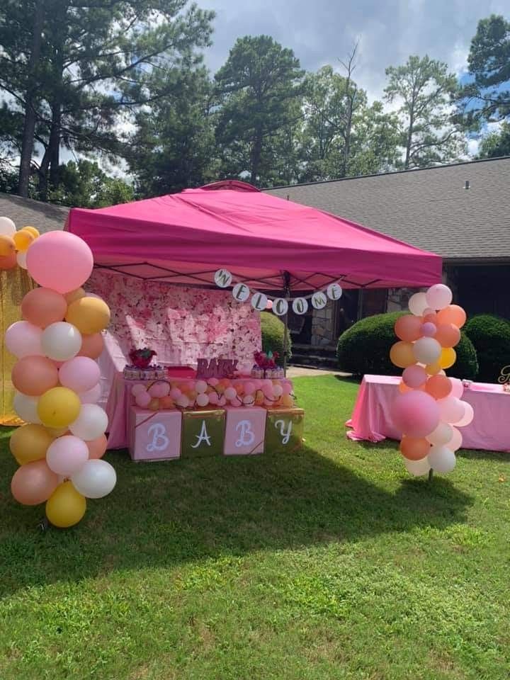 a pink and yellow baby shower with balloons on the grass near a table set up in front of a house