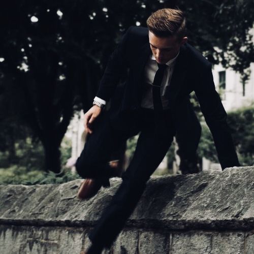 a man in a suit and tie doing a trick on a skateboard over a stone wall