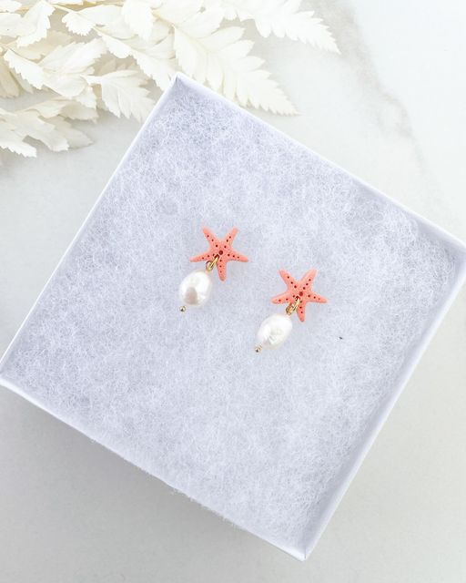 two starfish earrings with pearls on them sitting on a white box next to flowers