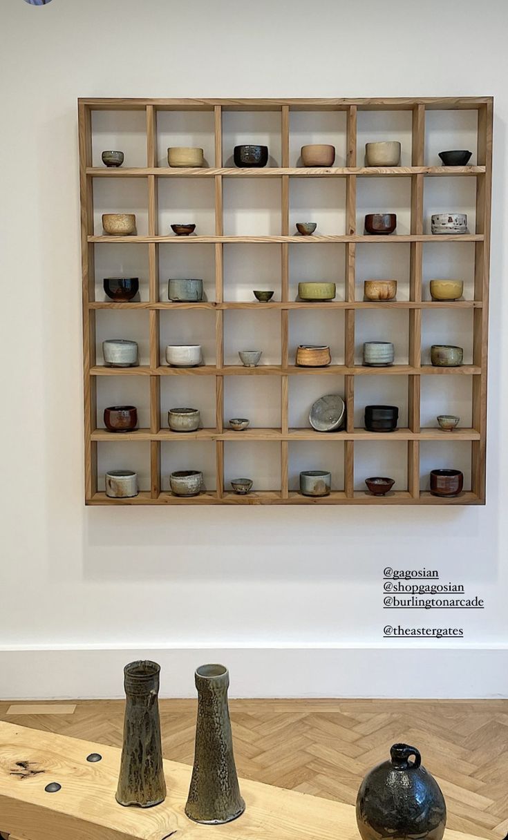 several vases and bowls are on display in a white walled room with wooden shelves