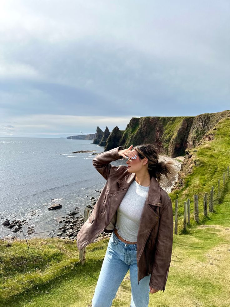 a woman standing on top of a lush green hillside next to the ocean with her hair blowing in the wind