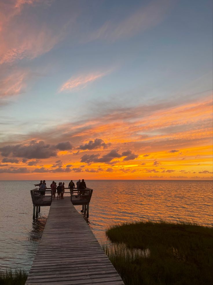 several people are sitting on benches at the end of a pier as the sun sets