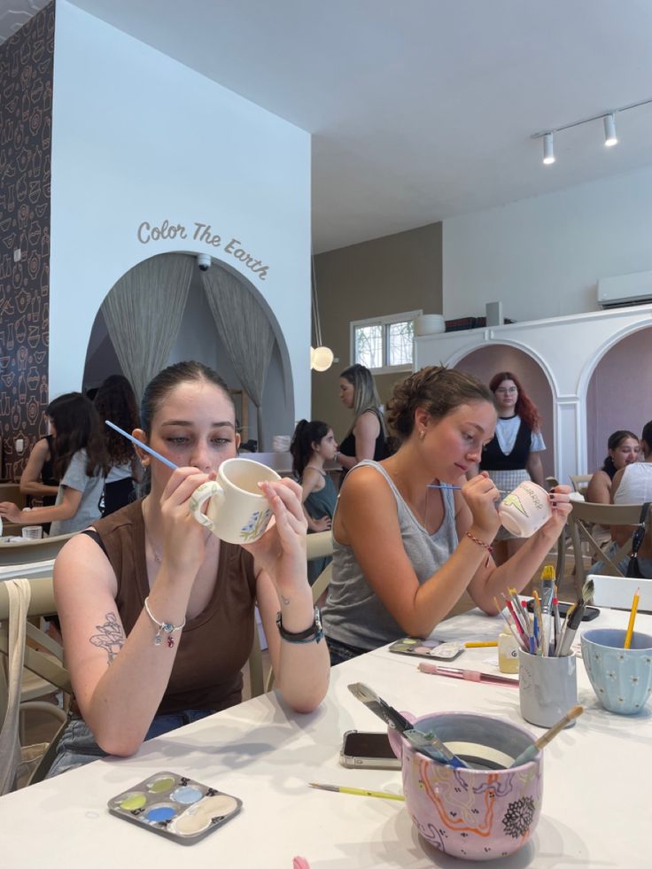 two women sitting at a table with cups and spoons in front of their mouths