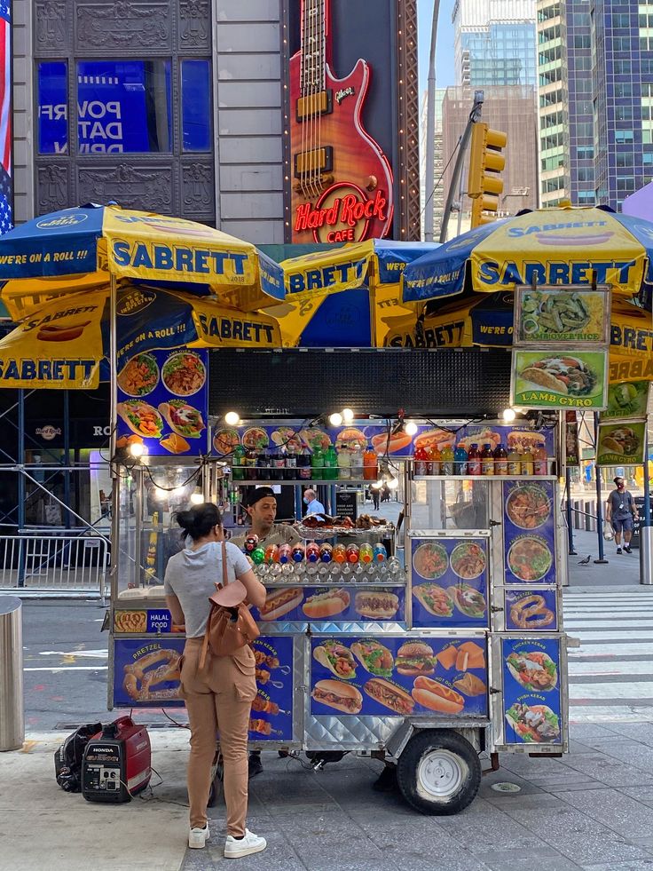 two people standing in front of a food cart on the side of the road near tall buildings