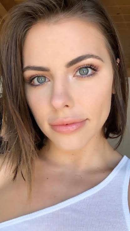 a woman with brown hair and blue eyes wearing a white tank top looking at the camera