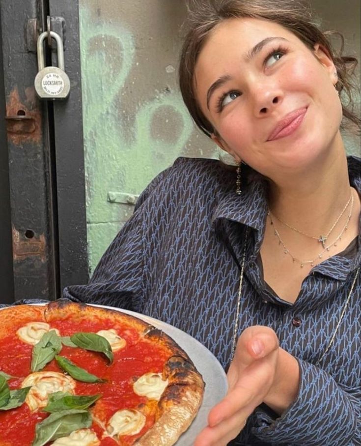 a woman holding a pizza with basil on it and looking up at the camera while sitting in front of a door