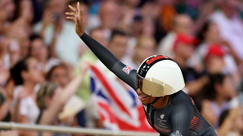 Kadeena Cox celebrating her gold medal win at the Stade de France veledrome, riding on her bike smiling with one arm in the air