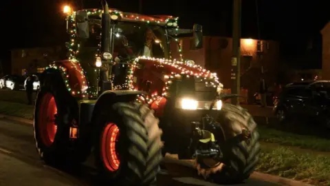 ANDREW MUTIMER A red tractor decorated with white lights is travelling through a darkly lit village street  