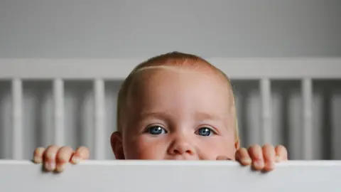 A baby with short reddish-blond hair and blue eyes lifts itself up to peek over the side of its cot