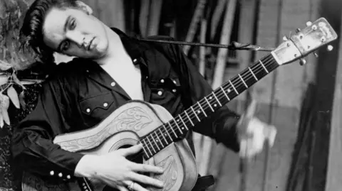 Elvis Presley poses for a portrait holding an acoustic guitar in 1956. He is wearing a dark-coloured shirt and is resting his head against a tree.