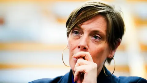 Jess Phillips wears large gold hoops, a blue blazer and holds her hand to her mouth. 