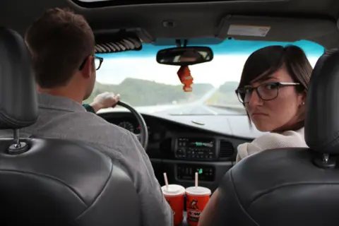 Shai Shotts A woman in the passenger seat looks behind her while a man drives the car