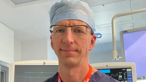 Leo Krivski looks at the camera, he is in a surgery theatre room with screens showing medical date behind him and wears blue scrubs. He has a red lanyard around his neck, dark blue scrubs and a pale blue hat on, with thin black wire framed glasses.