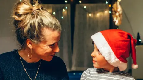 A woman and child smile at each other. The woman has her blonde hair tied up in a bun and is wearing a dark top and necklace. The child is wearing a Father Christmas hat that is too big for them. Behind the pair, Christmas lights shine.