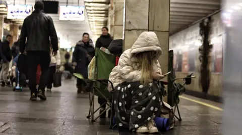 A woman sitting on a chair with the hood of her cream-coloured coat covering her face and a spotted blanket while she is on her mobile phone. Behind her, there are multiple people walking in the subway in Kyiv, where they hid during a Russian attack on 28 November, 2024