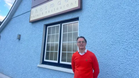 Mr Roberts stands outside the Plough Hotel and Restaurant in Rhosmaen, Llandeilo. He is wearing an orange jumper over a white shirt. 