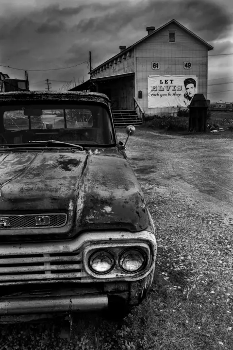 Geoff Watson An old car is parked in front of a sign with a picture of Elvis