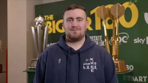 Luke Littler wearing a navy blue hoodie with the word "Nuke Luke" written on standing in front of his trophy.