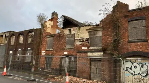 A two-storey derelict building with fencing around it. The red brick building has windows boarded up and the roof has caved in. There is grafitti on some of the boarded sections.