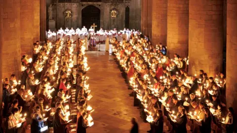 A large group of people singing inside Gloucester Cathedral during the candlelit service to mark the start of Advent. Many people can be seen either side of the main aisle each holding their own candle, while the choir can be seen in the distance