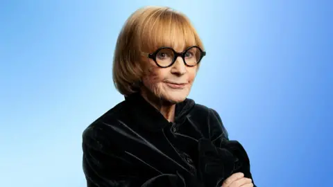 A portrait image of Anne Robinson, wearing black round glasses and a black coat. She has her arms folded