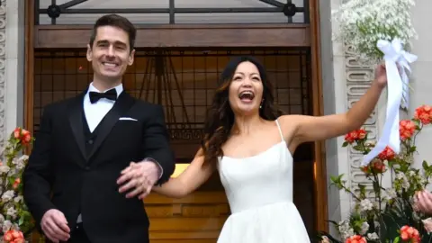 A bride and groom come out of a registry office having just got married. The bride has long dark hair and wears a white dress with spaghetti straps and shoulders mostly exposed, and holds her bouquet of flowers aloft. The groom is wearing a traditional black dinner jacket and has neat, dark hair. The newlywed couple are holding hands and smiling.