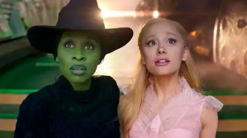 Cynthia Erivo as Elphaba and Ariane Grande as Galinda in the film Wicked. Erivo has green skin, and wears a black dress and witch's hat. Grande has chest-length blond hair and wears a pink dress. They both stare at something off-camera. Behind them, a light is shining.