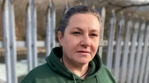 Sharon Elward, local organiser for Pontypridd foodbank stares at the camera. She is standing in front of the river Taff in Pontypridd, wearing a green hoody. Metal railings can be seen behind her, and beyond them the river.  