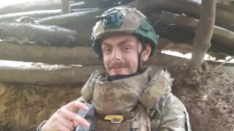 Callum Tindal-Draper wearing military camouflage clothing while stood in front of a set of logs in Ukraine. 