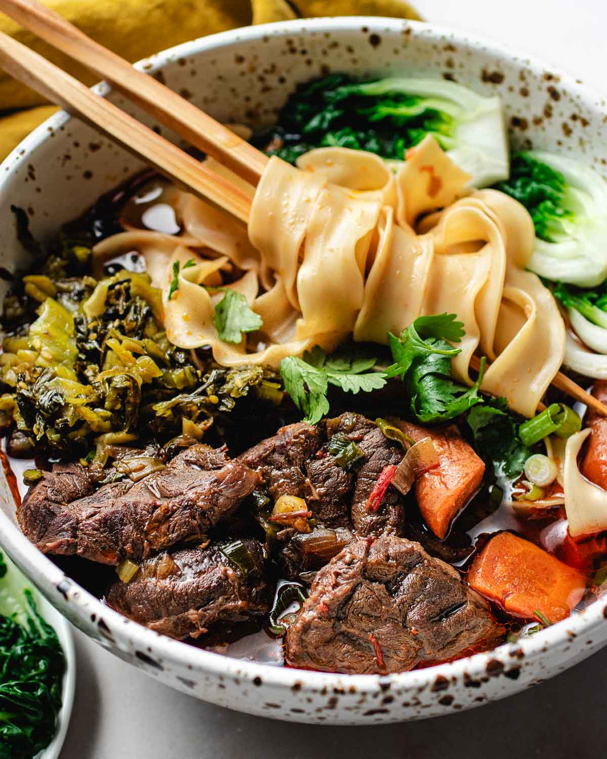 Feature image shows a big bowl of Taiwanese beef noodle soup served with bok choy and pickled mustard greens in the bowl with chopsticks.