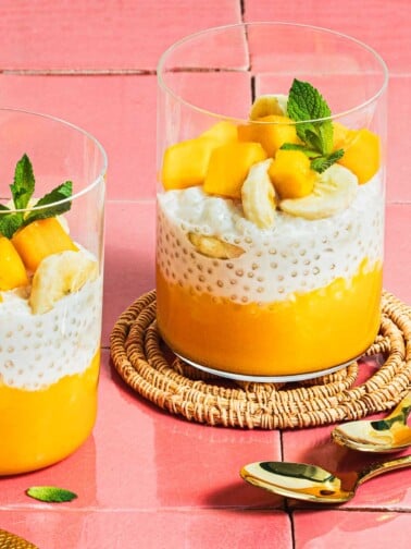 Image shows 2 glasswares filled with mango puree, sago pudding, and chopped mango and mint leaves served as a dessert.