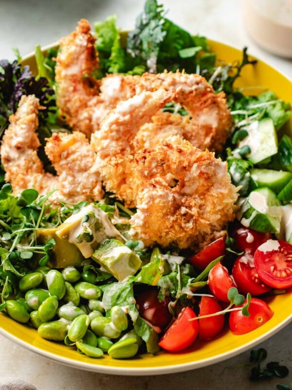 Image shows a side close shot with crispy bang bang shrimp on top of a bowl of salad greens and vegetables, served in a yellow color bowl.