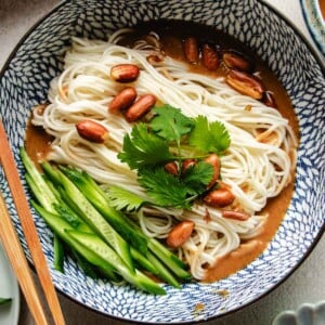 An overhead shot image shows a blue and white color bowl with boiled cold noodles inside with Peanut butter sauce and veggies on the side.